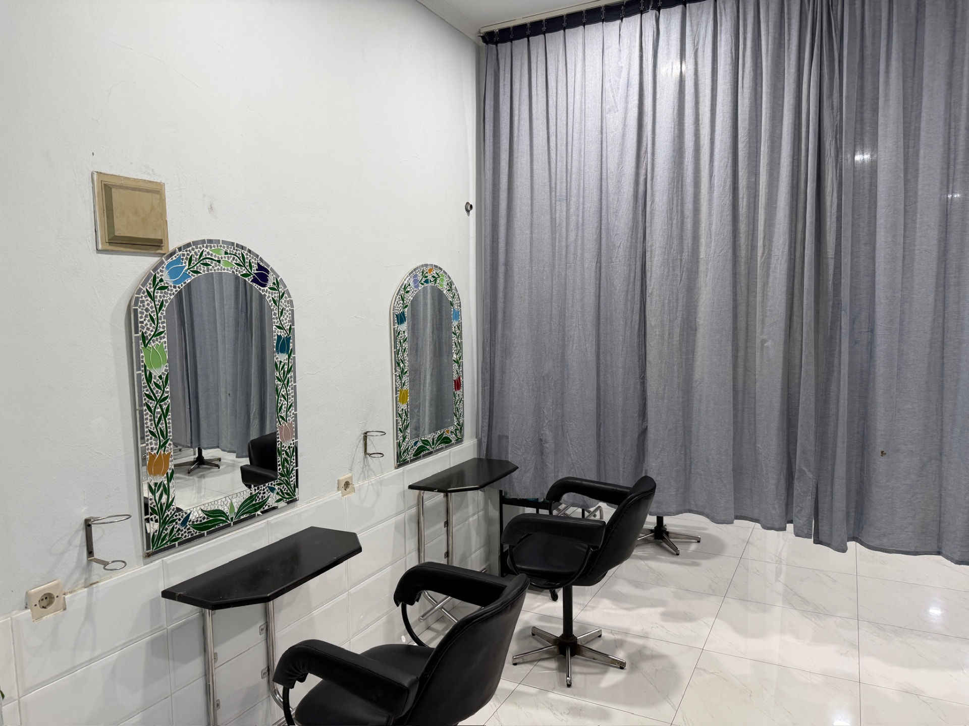 Ruang styling hijab pribadi di Yucca Salon dengan dua cermin lengkung mosaic, kursi salon hitam, tirai abu-abu, dan dinding keramik putih, Bandung